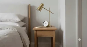 A narrow nightstand styled with a contemporary brass reading lamp, a ceramic coaster, and a minimalist alarm clock.